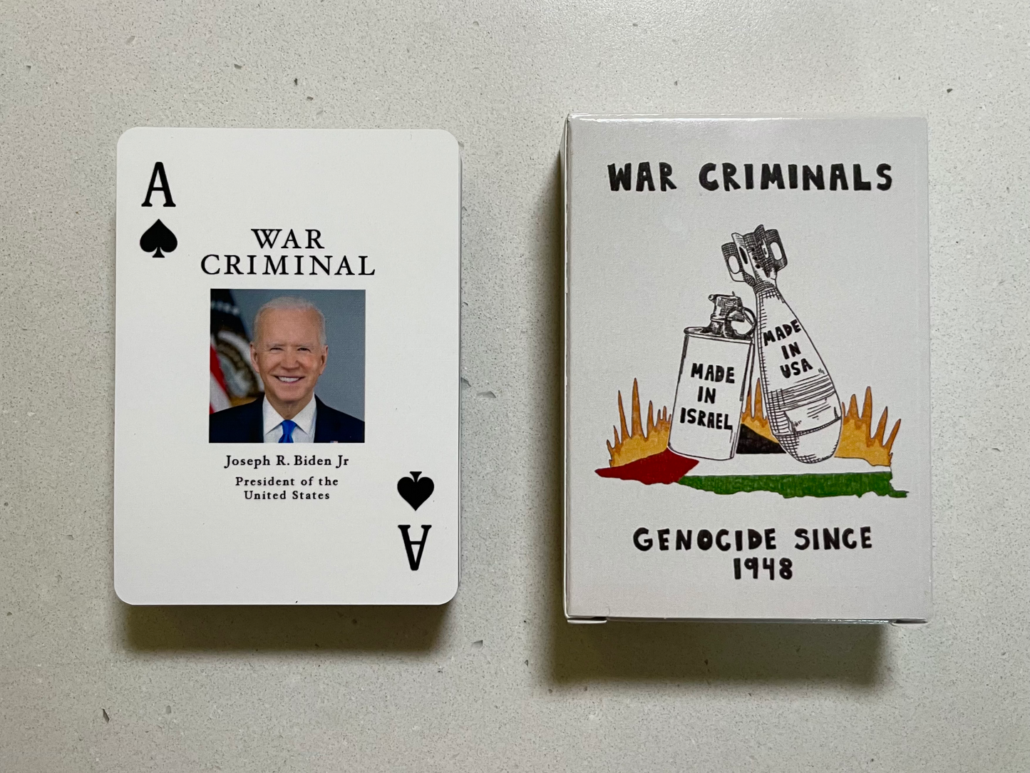 A photo of playing cards box and the Ace of Spades laid side by side on a white stone background. The Ace of Spades centers a photo of Joseph R. Biden, Jr and includes the title “War Criminal" above the photo and his name and professional title below the photo. The War Criminals Playing Cards box includes an illustration of a tear gas canister and a bomb, the former inscribed with "Made in Israel" and the latter inscribed with "Made in USA". 
